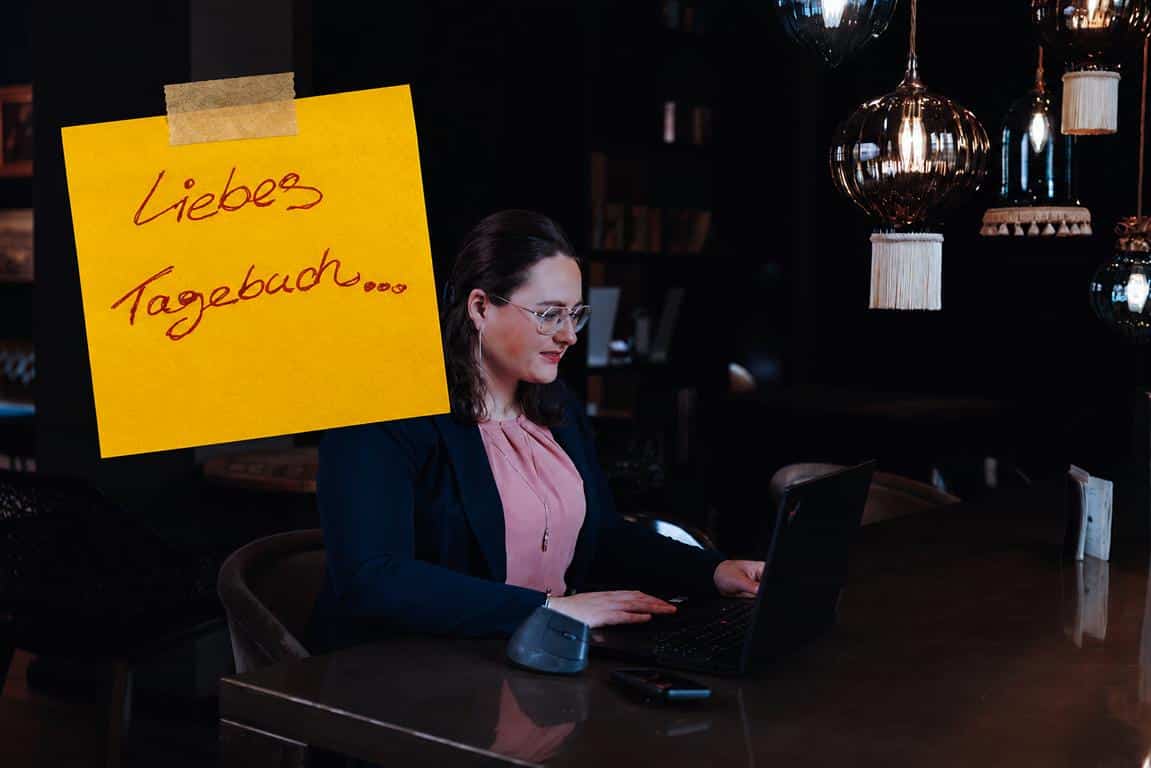 Antonia Hillmann sitzt mit Laptop in einer dunklen Lounge-Atmosphäre, trägt einen dunkelblauen Blazer und eine rosafarbene Bluse. Über dem Bild klebt ein gelber Notizzettel mit der handgeschriebenen Aufschrift „Liebes Tagebuch…"